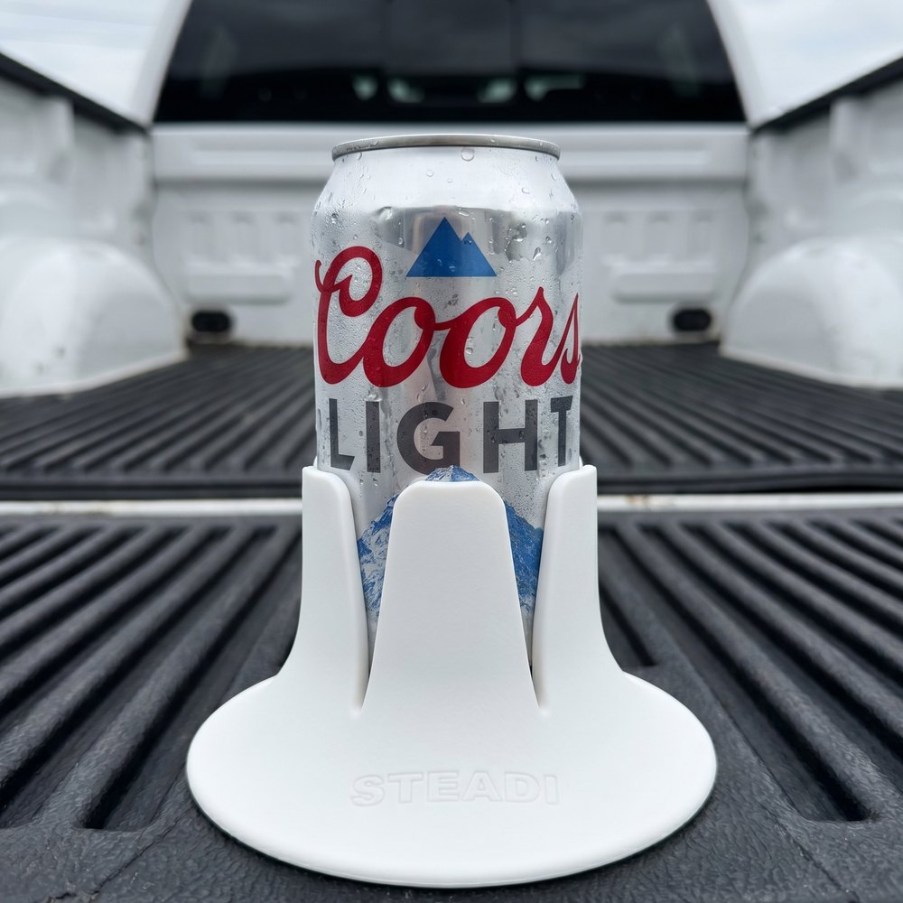 A cold Coors Light can, beaded with condensation, standing in a white Steadi on the ribbed black bed mat of a pickup truck.